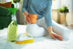 Nahaufnahme einer Frau in gestreiftem T-Shirt und weißer Hose mit Sprühflasche mit grünem Reinigungsmittel, gelber Bürste und einer Tasse Kaffee, die an sonnigem Tag den Teppich im Haus reinigt